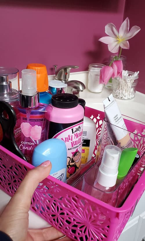 closeup photo of hand and pink plastic basket of toiletries: body spray, deodorant, mouthwash, powder, makeup. Sink in background with Q-tips and pink tulips
